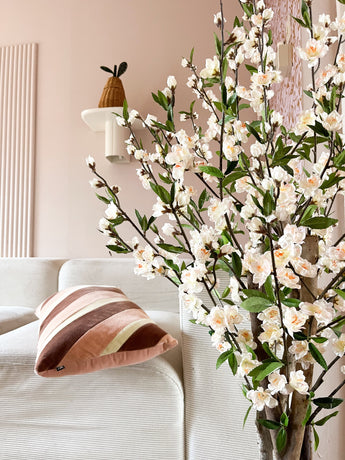 Een gezellige woonkamer met een witte bank, een gestreept roze en bruin kussen en de Kunstplant Kersenbloesem 120 cm crème bloeiende tak op de voorgrond. Een potplant rust op een wandplank tegen een lichtroze muur.