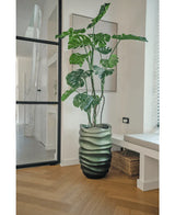 De Kunstplant Monstera 150 cm, met zijn grote groene bladeren, staat in een pot met golvend patroon op een houten vloer in een moderne, lichte kamer met een glazen deur, ingelijste kunst en een rieten mand in de buurt.