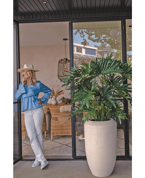 En kvinna i blå blus, vita byxor och hatt skrattar åt en glasdörr bredvid växten Artificiell Philodendron 125 cm i en beige kruka. Korgstolar och inredning syns i hela rummet.