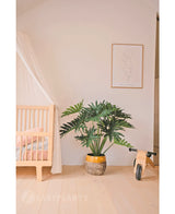 Een gezellige, minimalistische kinderkamer met links een houten wieg en luifel, in het midden een Kunstplant Philodendron UV 90 cm als pronkstuk, rechts een houten speelgoedfiets en daarboven een eenvoudige lijnkunstprint.