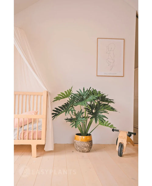 Ett mysigt, minimalistiskt barnrum med en trävagga och baldakin till vänster, en Artificiell Philodendron UV 90 cm växt i mitten som ett utställningsexempel, en leksakscykel i trä till höger och ett enkelt linjekonsttryck ovanför.