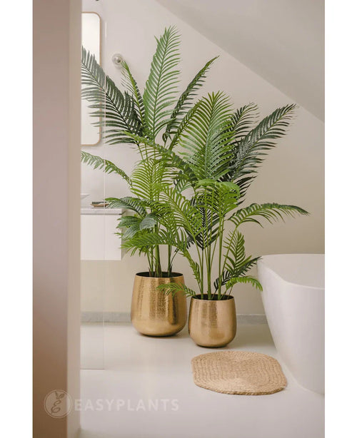 En konstgjord växt Phoenix 180 cm med frodiga gröna blad i en glänsande guldkruka står bredvid ett vitt badkar och en beige vävd matta i ett modernt, minimalistiskt badrum, kompletterat med en spegel och ett fönster för en fridfull look.