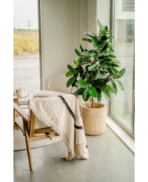 En trästol med en krämfärgad matta står bredvid Konstgummiväxten 150 cm i snygga krukor nära höga fönster, där naturligt ljus och ett ljusgrått golv förstärker den ljusa, fridfulla atmosfären i rummet.