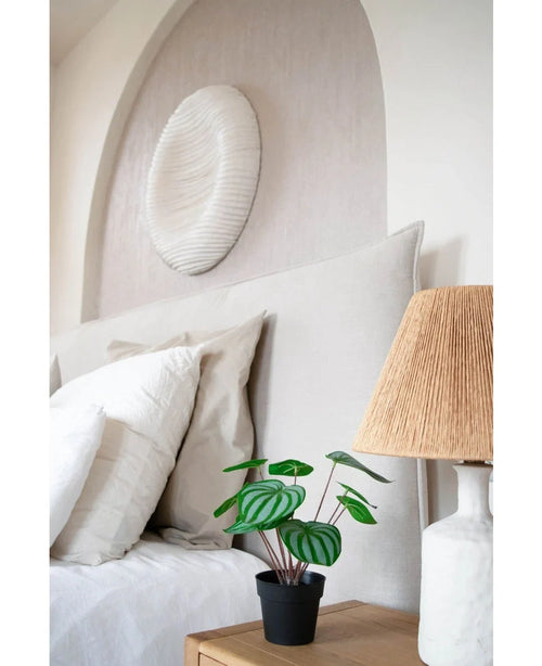 En prydligt bäddad säng med vita kuddar och sängkläder står bredvid ett sängbord i trä med den konstgjorda vattenmelonplantan 29 cm och en lampa med vävd skärm; ovanför sänggaveln hänger en vit texturerad väggskulptur.