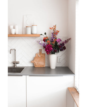 Ett modernt vitt kök med en grå bänkskiva, ett handfat, en skärbräda i trä och en vit vas fylld med konstgjord blomhortensia 55 cm mörkrosa. Vita koppar och krukor står på en trähylla ovanför disken.