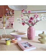 Ett modernt rum har ett dukat bord med konstgjord gren Magnolia liten blommig 89 cm rosa i rosa vas, pastellfärgade ljusstakar, skålar, koppar, en grön skulptural vas, en rosa bok och blommiga konstverk på väggen.