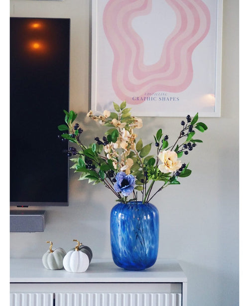 En blå glasvas med en artificiell grenbär 76 cm lila konstgjord blomma står på ett vitt skåp. Bredvid står två vita pumpor med guldstjälkar. På väggen bakom den finns en TV och ett abstrakt rosa konsttryck.