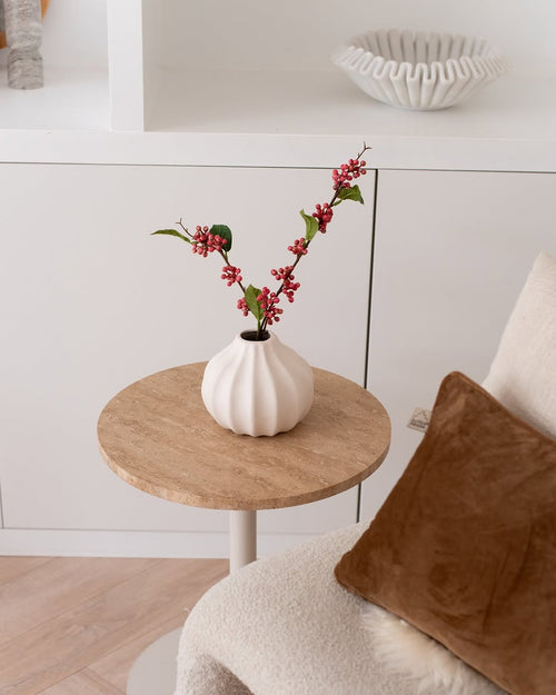 Artificiella grenbär 64 cm rosa - en vit vas fylld med rosa konstgjorda bärgrenar och gröna blad - står på ett litet runt bord bredvid en beige stol med en brun kudde i ett mysigt minimalistiskt rum.
