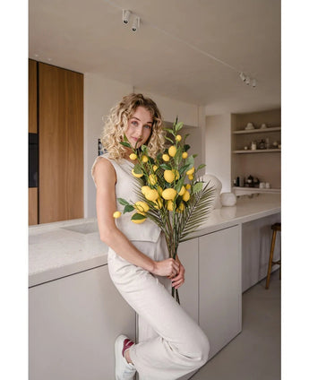En kvinna med lockigt blont hår, klädd i en krämfärgad outfit, lutar sig mot en disk och håller buketten 'Artificiell Citrongren 104 cm gul', dekorerad med frodig grönska. Det moderna köket är inrett i neutrala färger och har trädetaljer.