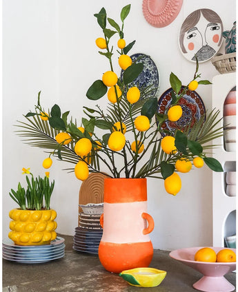 Ett ljust rum med en hög vas full av citroner och gröna löv, där den slående Kunsttak Lemon 104 cm gul intar en framträdande plats i arrangemanget, omgivet av färgglada keramikskålar, en skål med citroner och dekorativa tallrikar på väggen - skapar en glad, eklektisk look.