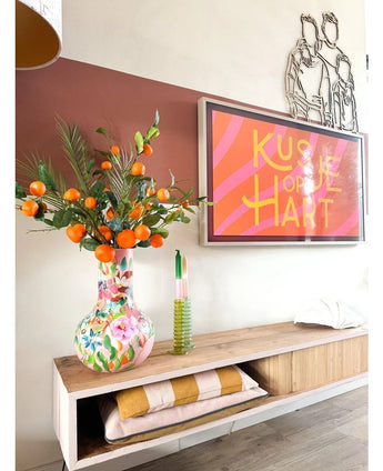 På en trähylla står en färgglad vas innehållande Art Branch Mandarin 93 cm orange, bredvid ett inramat konstverk med rosa ränder där det står ”Kissje op je Hart”. På hyllan finns också ett grönt ljus och vikta filtar.