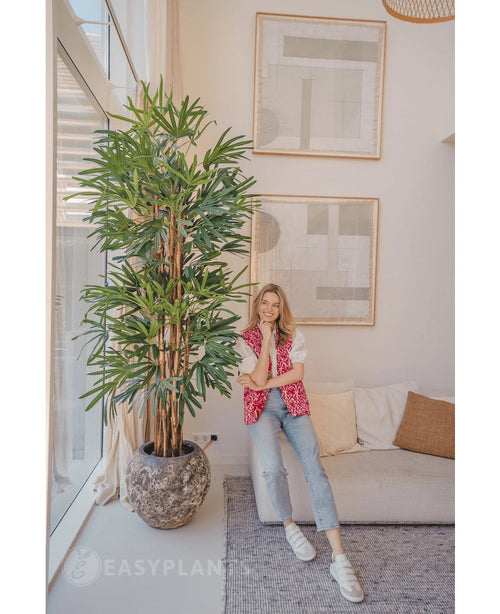En kvinna i en rosa mönstrad kofta och jeans ler på en beige soffa bredvid Lady Artificial Palm 210 cm i ett ljust, modernt vardagsrum med abstrakt konst på väggen.