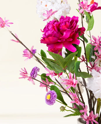 Het Zijden kunstbloemen boeket Pink Reverie heeft een grote magenta pioenroos, kleine roze bloemen, paarse madeliefachtige bloemen en groene bladeren in een vaas-een romantisch boeket tegen een lichte achtergrond.