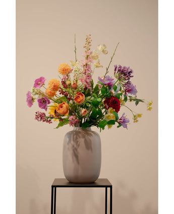 Een beige vaas met Ranonkel kunsttak x5 Real Touch oranje 62cm kunstmatige oranje ranonkels staat op een zwart metalen standaard tegen een effen lichte achtergrond.