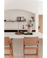 Een minimalistische keuken heeft een wit eiland met twee houten stoelen en de Vaas Artic Beige Ø21 x H29 cm met roze en witte bloemen. Aan de muur op de achtergrond zijn planken met boeken en keramiek bevestigd.
