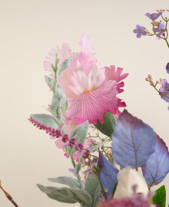 Een close-up van het Zijden kunstbloemen boeket Amethyst Romance, met een grote roze en witte bloem, zachtgroene bladeren, kleine paarse bloesems en blauw getint gebladerte tegen een neutrale achtergrond.