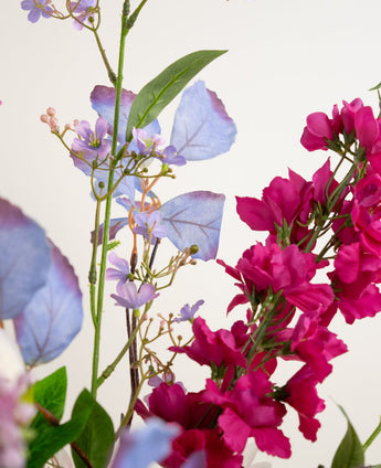 Close-up van het Zijden kunstbloemen boeket Amethyst Romance, met levendige magenta en paarse bloemen met lichtblauwe bladeren en groene stelen op een effen witte achtergrond.